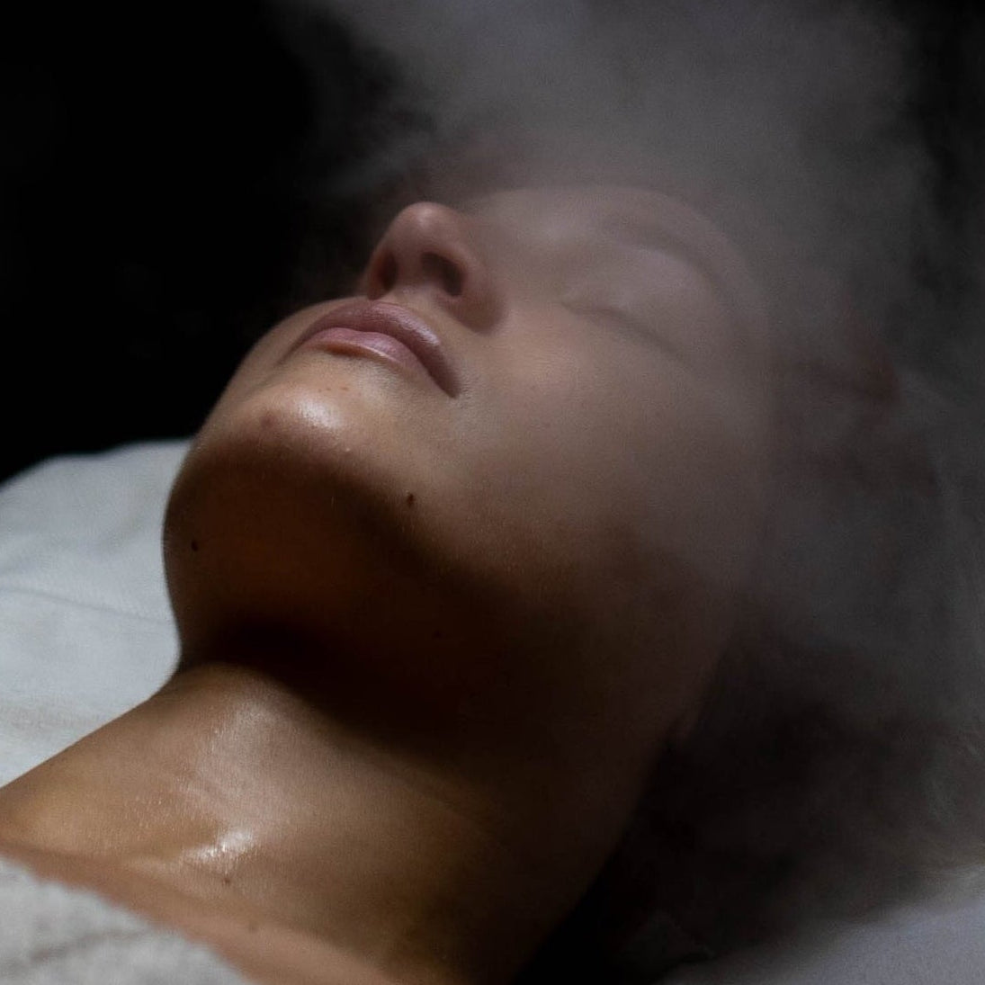 Person receiving a facial treatment with a light and steam spray in a dimly lit room.