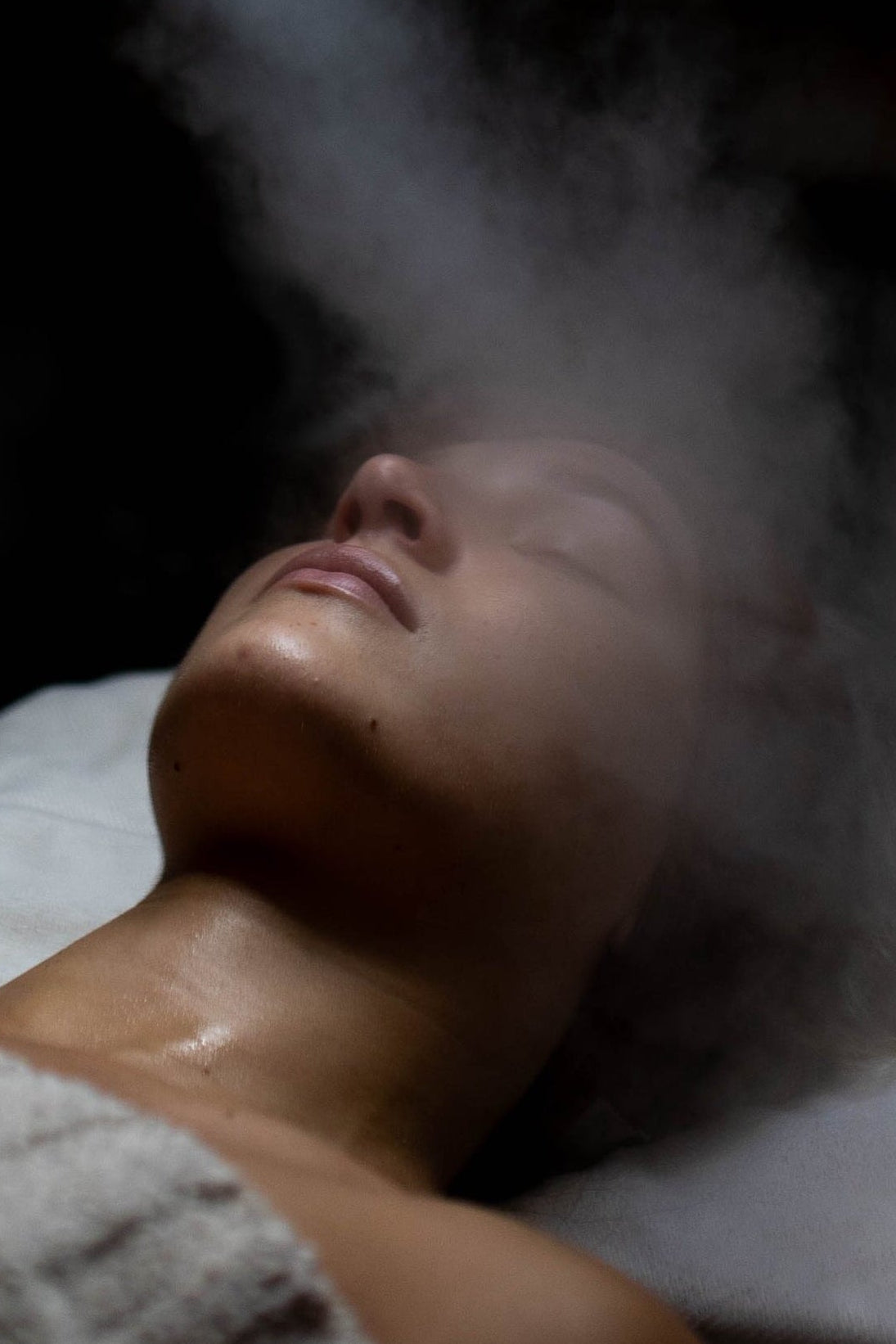 Person receiving a facial treatment with a light and steam spray in a dimly lit room.